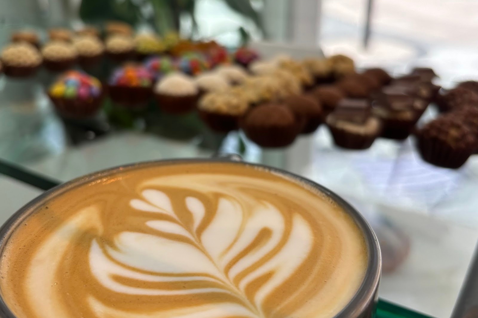 A coffee with rosetta latte art on a glass counter, with a case of brigadeiros behind it.